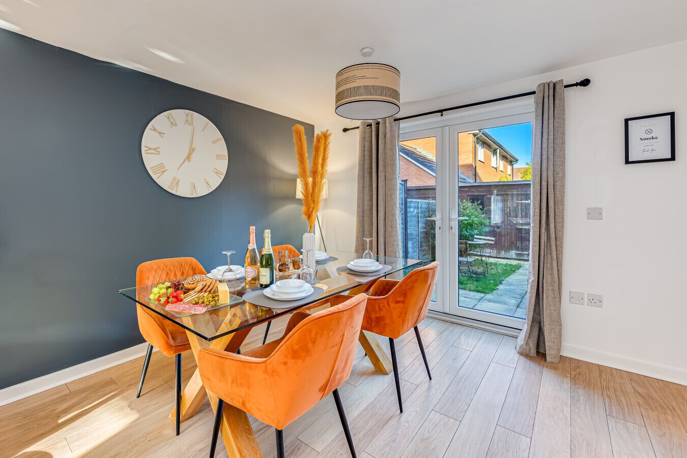 Een moderne eetruimte met een glazen tafel, oranje stoelen en een grote wandklok.