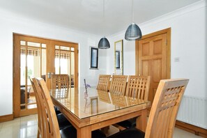 Een eetgedeelte met een tafel met glazen blad, houten stoelen, hanglampen en een houten deur.