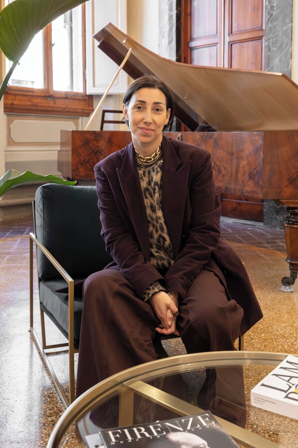 A person seated in a modern room with a piano, a glass table with a book, and a large plant. - Maggio Living (Florence)