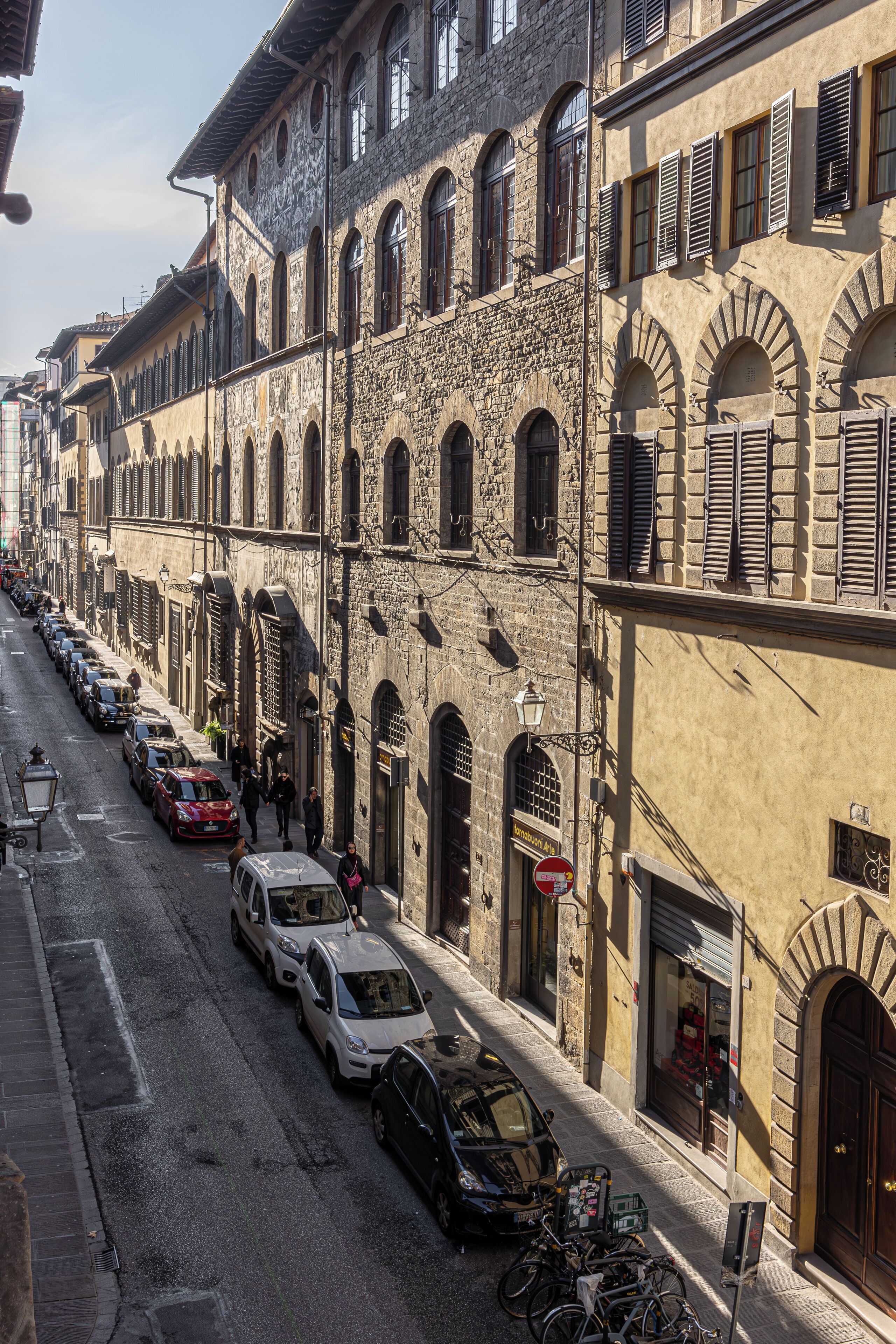 Una strada fiancheggiata da edifici storici in pietra, auto parcheggiate e biciclette.