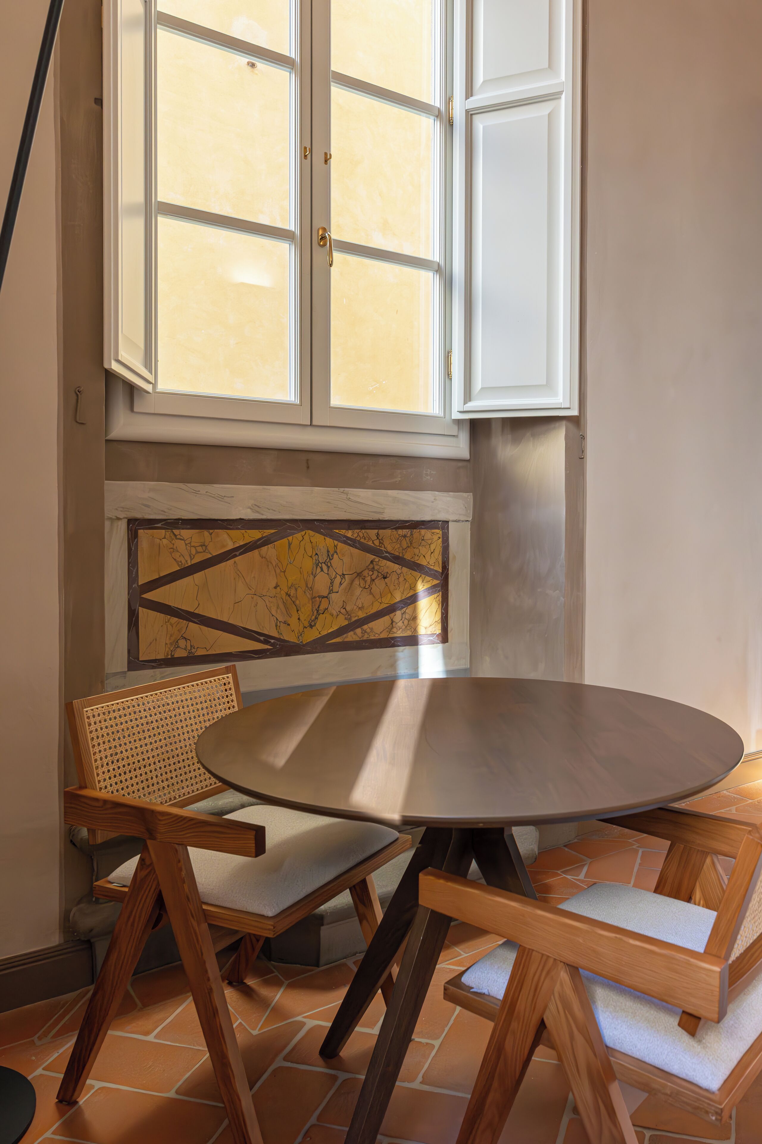 A dining area with a round table and two wooden chairs, a window with white shutters, and a marble-patterned wall.