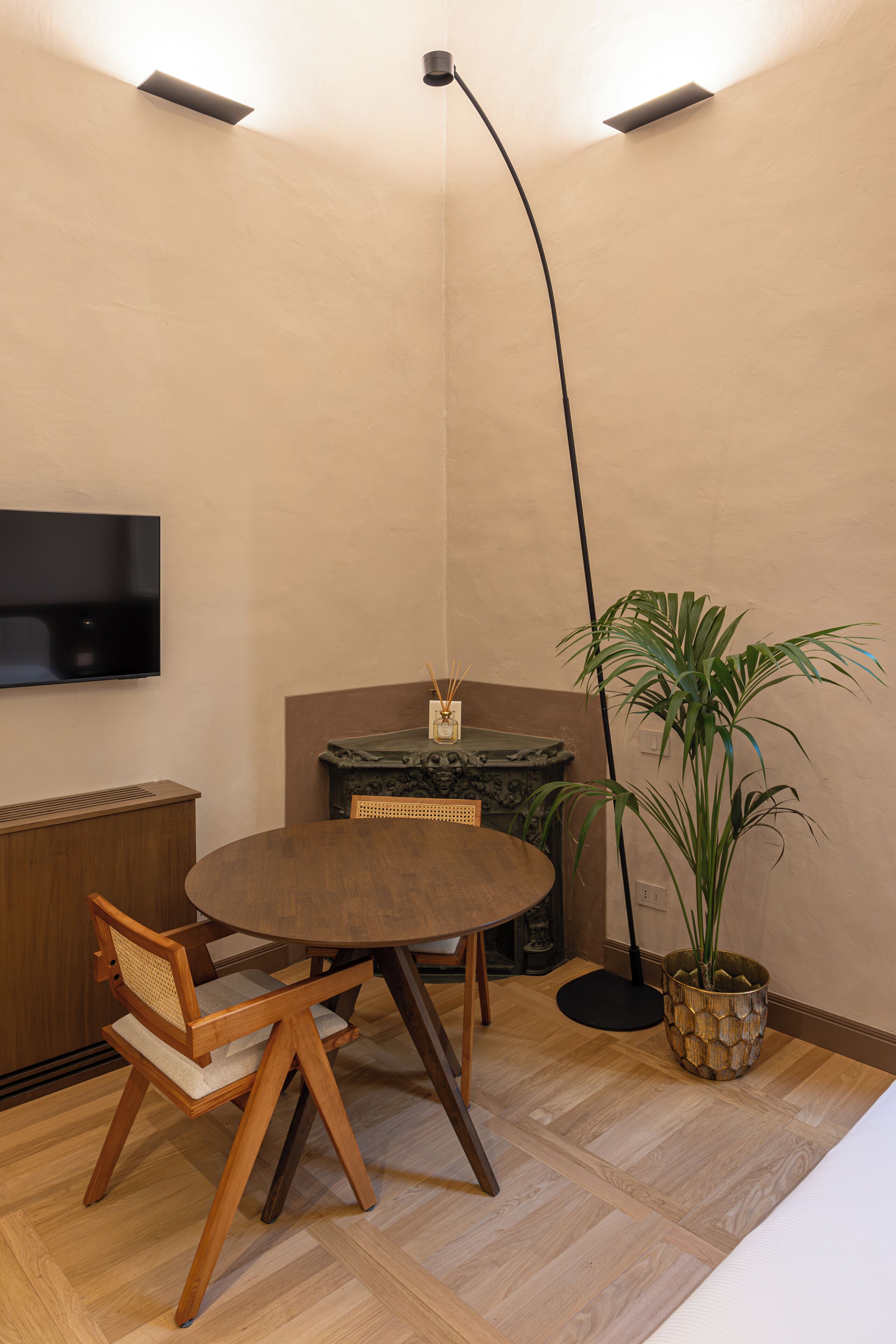 A small, cozy room with a round wooden table, two chairs, a television mounted on the wall, a floor lamp, a potted plant, and a marble fireplace.