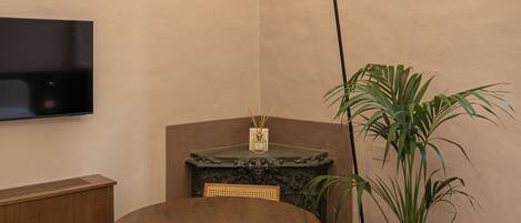 A small, cozy room with a round wooden table, two chairs, a television mounted on the wall, a floor lamp, a potted plant, and a marble fireplace.