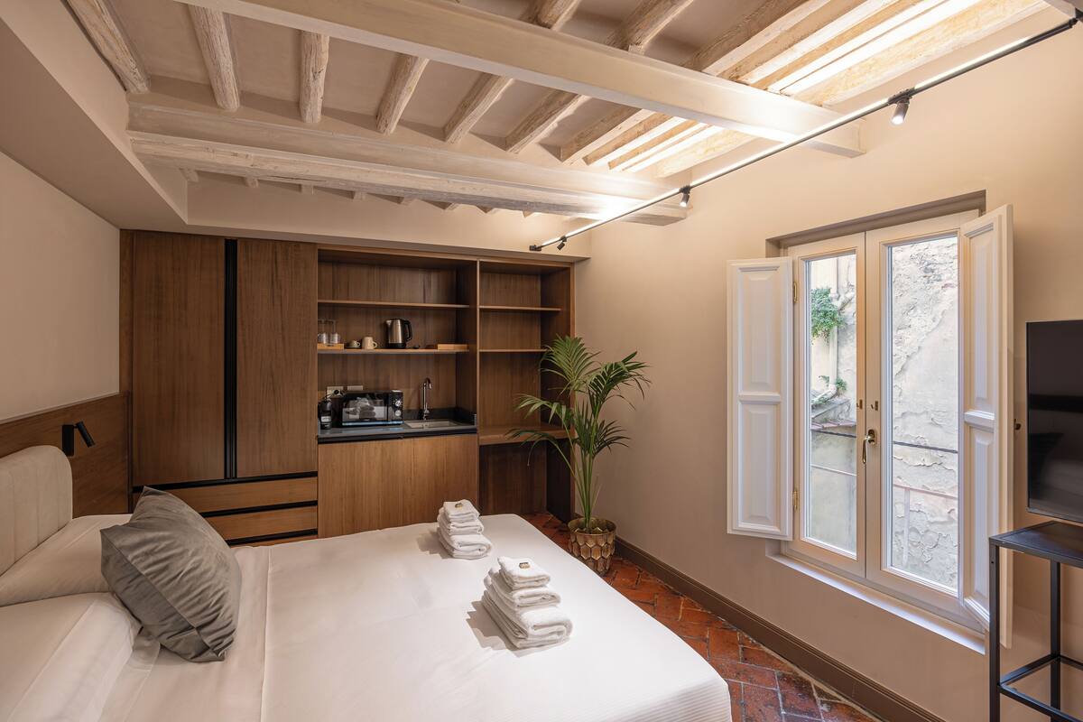 A hotel room with a bed, a built-in kitchen area, a TV, and a large window with a view of the outside.