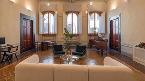 A spacious lobby with a high ceiling, large windows, a piano, and a seating area with a coffee table. - Maggio Living (Florence)
