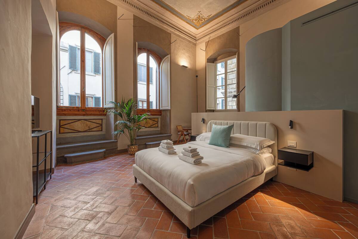 A bedroom with a large bed, a potted plant, and a view of the outside through arched windows.