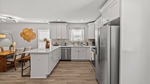 A modern kitchen with white cabinetry, a central island, stainless steel appliances, and a dining area with a wooden table and chairs. - Modern 4BR Retreat near Downtown Concord (Concord)