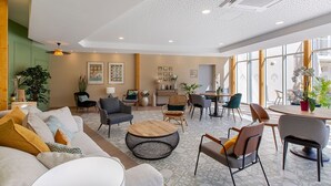 A spacious lobby with a mix of seating areas, including sofas and chairs, a coffee table, and a dining table with chairs. There are plants and artwork on the walls, and the ceiling has a perforated design with recessed lighting. - Résidence de Bourges (Bourges)
