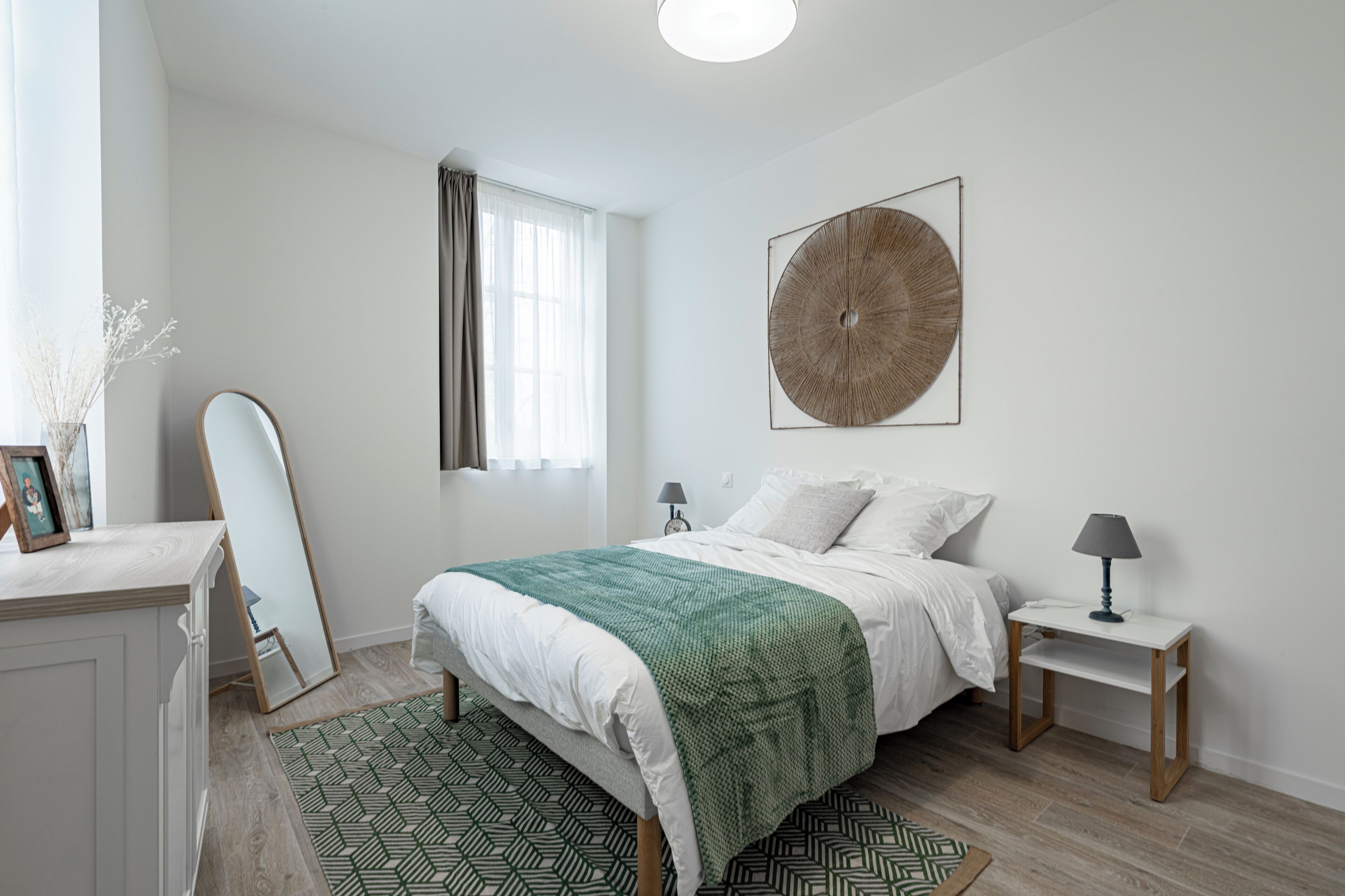 Une chambre à coucher comprenant un lit, des tables de chevet, un miroir, une œuvre d’art murale et une fenêtre avec des rideaux.