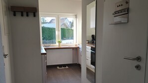 Un couloir étroit mène à une cuisine dotée d’une fenêtre, d’un radiateur et d’une porte ornée d’un miroir et d’une petite étagère.