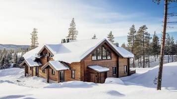 En stuga av trä med ett snötäckt tak, omgiven av träd och ett snöigt landskap.