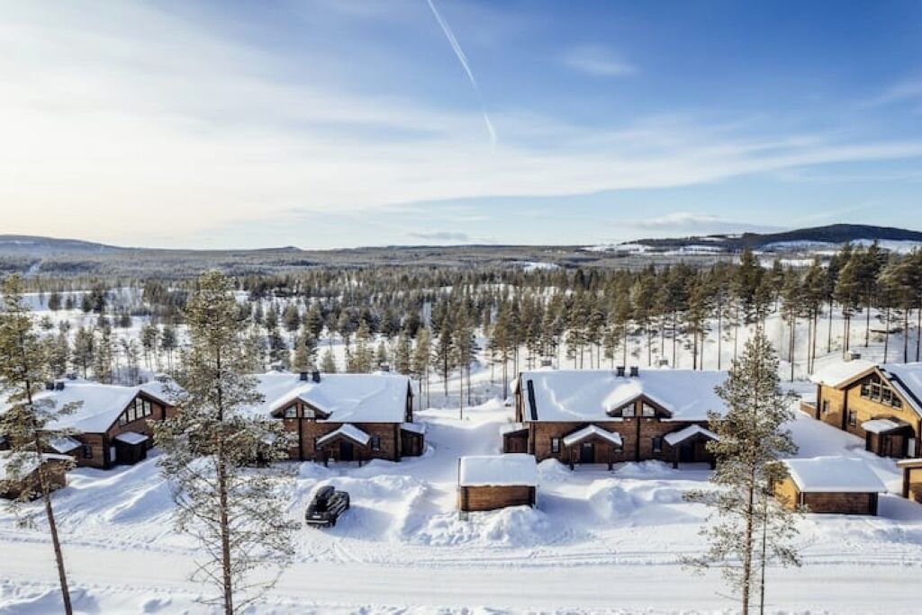 A snow-covered lodge with multiple cabins, surrounded by a forest and distant mountains.