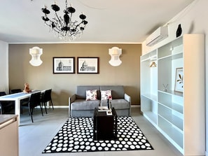 A modern living room with a grey sofa, a black coffee table, a chandelier, and a dining area with a white table and chairs.