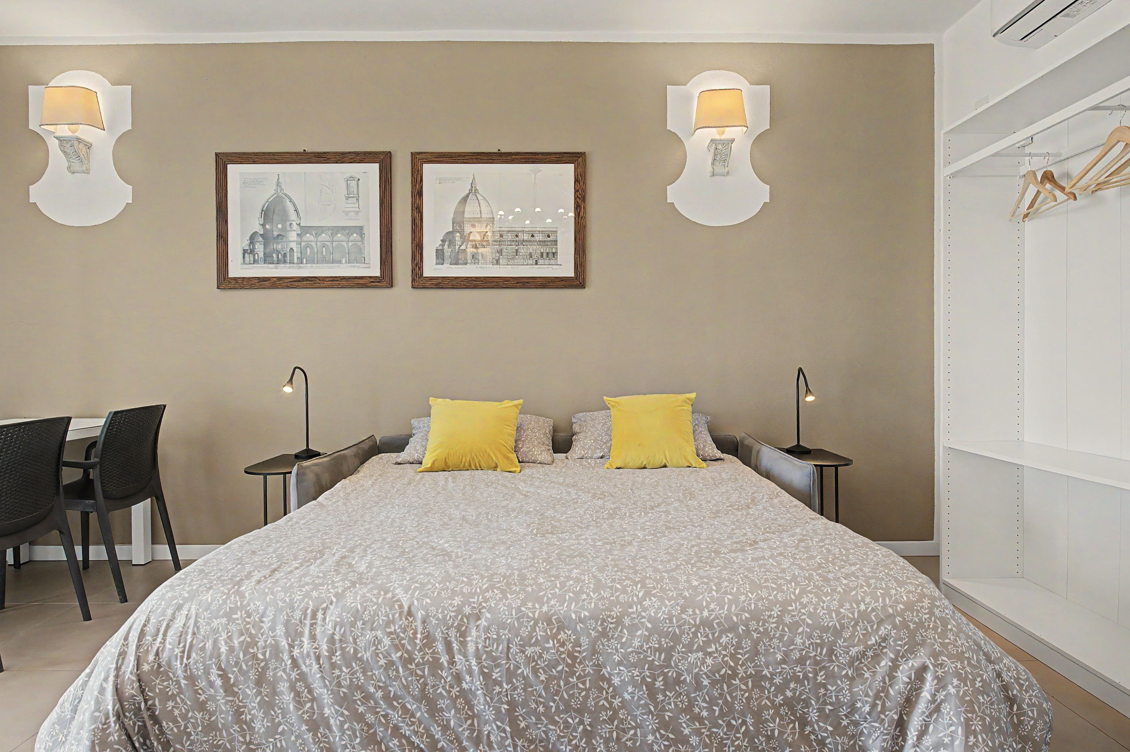A bedroom with a large bed, two bedside lamps, a desk with a chair, and two framed pictures on the wall.