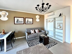 A modern living room with a grey sofa, a dining table with chairs, a chandelier, framed pictures on the wall, and a white closet with hanging space.