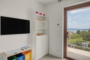 A room with a white wardrobe, a flat-screen TV mounted on the wall, and a window with a view of a landscape. - Panoramic Apartment with Lake View, Wi-Fi, Air Conditioning & Private Parking (Padenghe Sul Garda)
