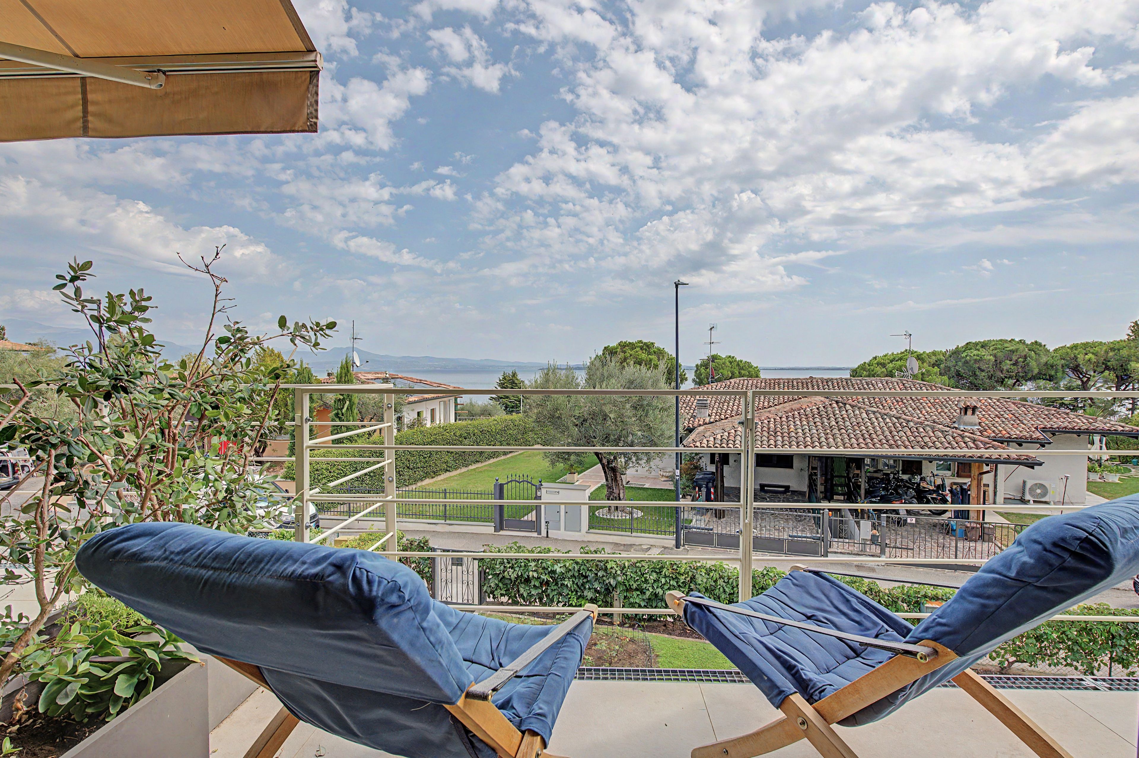 Un balcón con dos sillones de descanso, vistas despejadas a una zona residencial y un cielo parcialmente nublado.