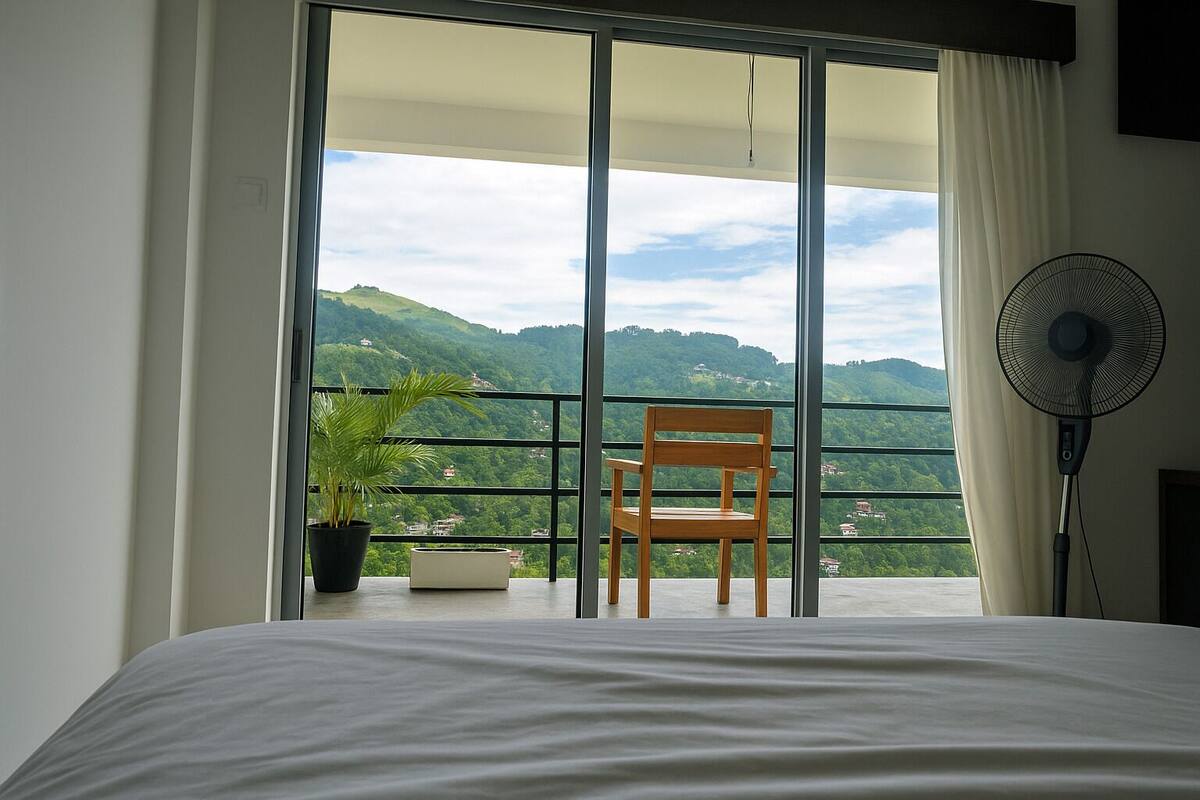A bedroom with a bed, a chair, a potted plant, and a balcony with a view of green hills.