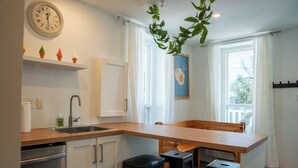 A modern kitchen with a wooden countertop, a sink, and a trash bin. There is a clock on the wall and a window with curtains.