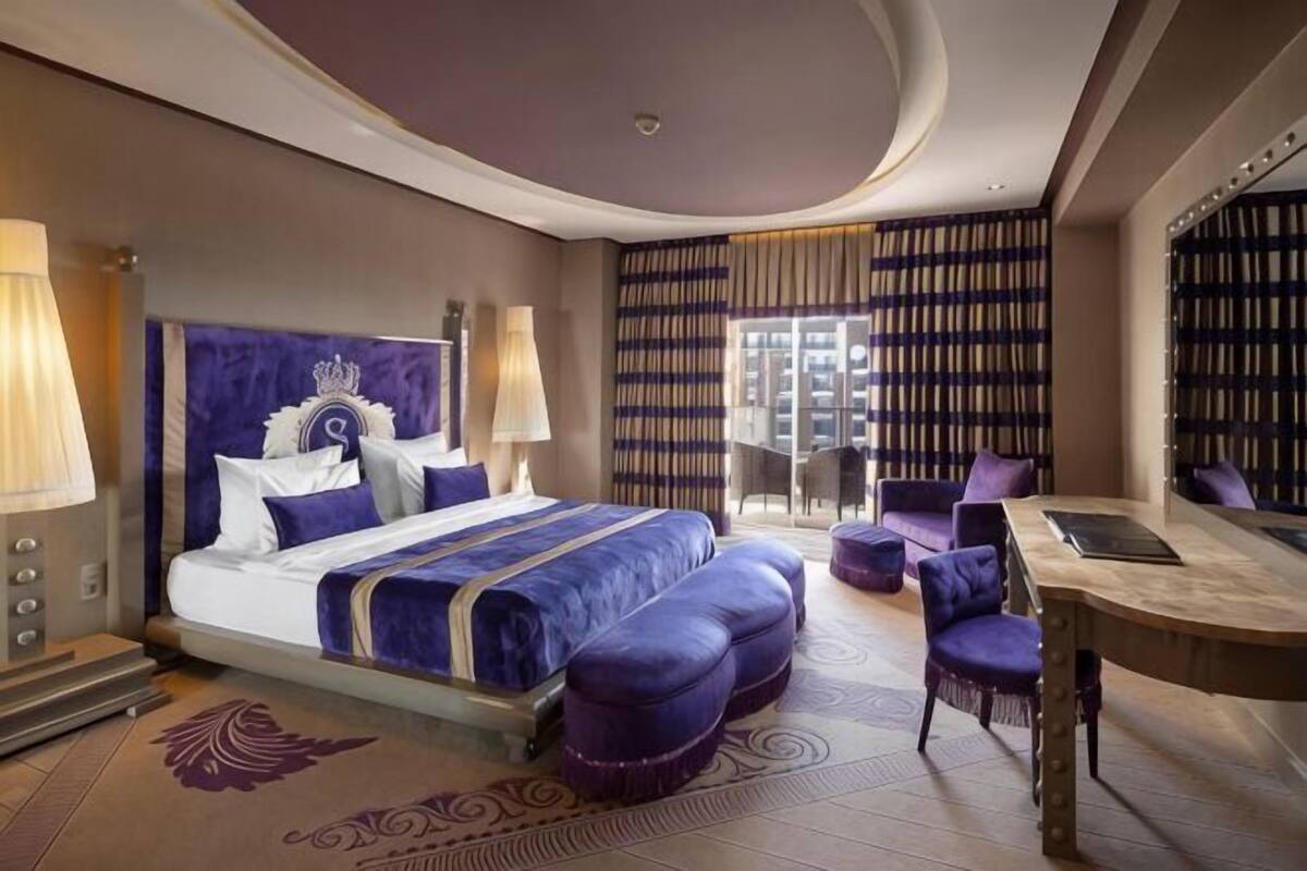 A hotel room with a large bed, a desk, and a chair. There is a bookshelf and a view of the city.