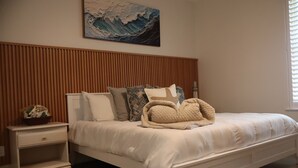Een slaapkamer met een houten lattenkap, een wit bed met meerdere kussens, een nachtkastje met een lamp en een kunstwerk van een golf aan de muur.