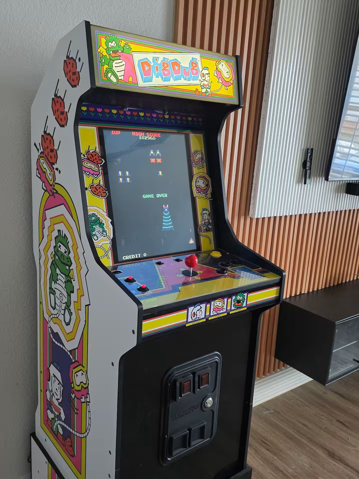 Een retro spelcomputer met een kleurrijk ontwerp, een scherm waarop een spel wordt getoond en een muntgleuf onderin.