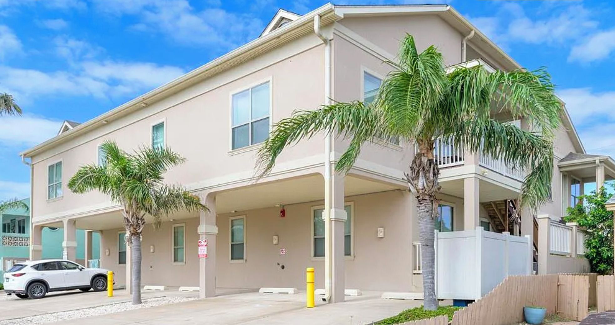 A two-story house with a balcony, a white car parked in front, and palm trees.