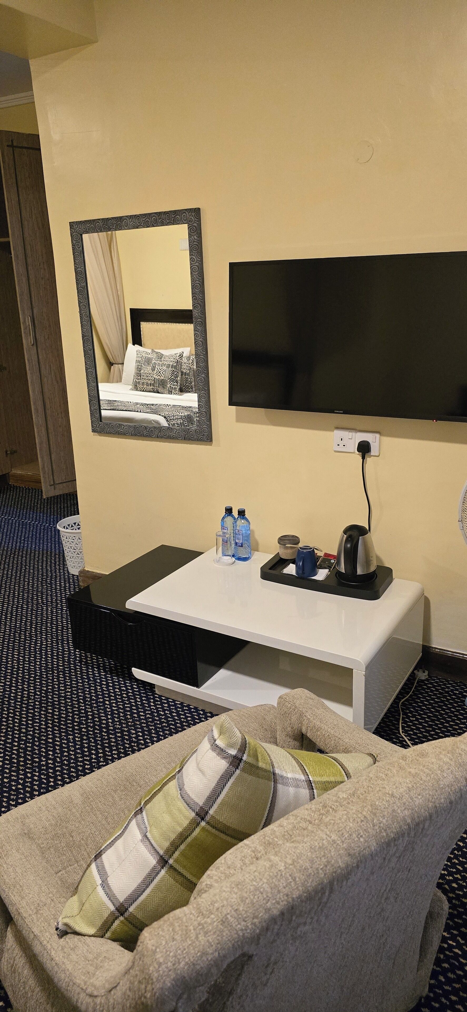 A hotel room with a bed, a bedside table with a tray of toiletries, a flat-screen TV mounted on the wall, and a patterned floor.