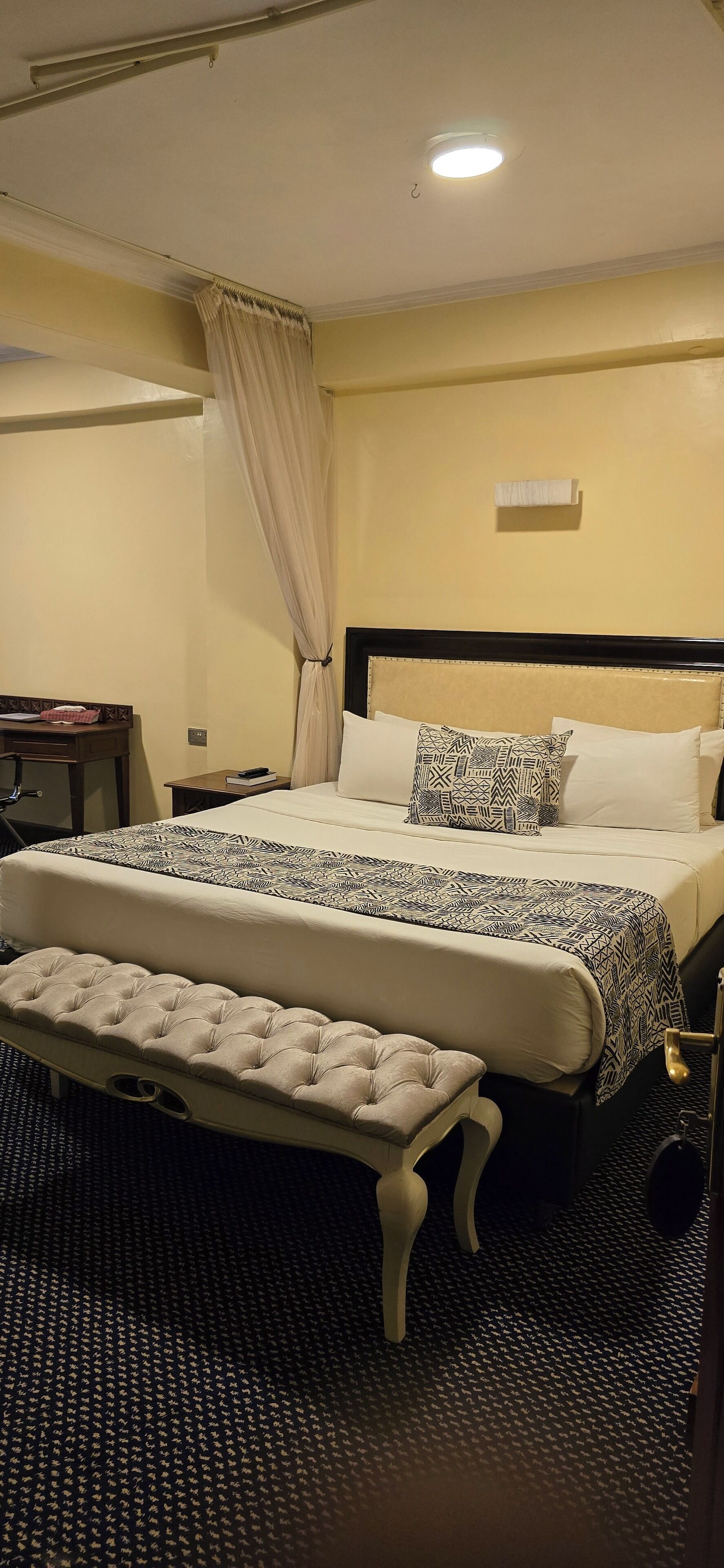 A hotel room with a bed, a bedside table, a chair, a bench, and a carpeted floor.