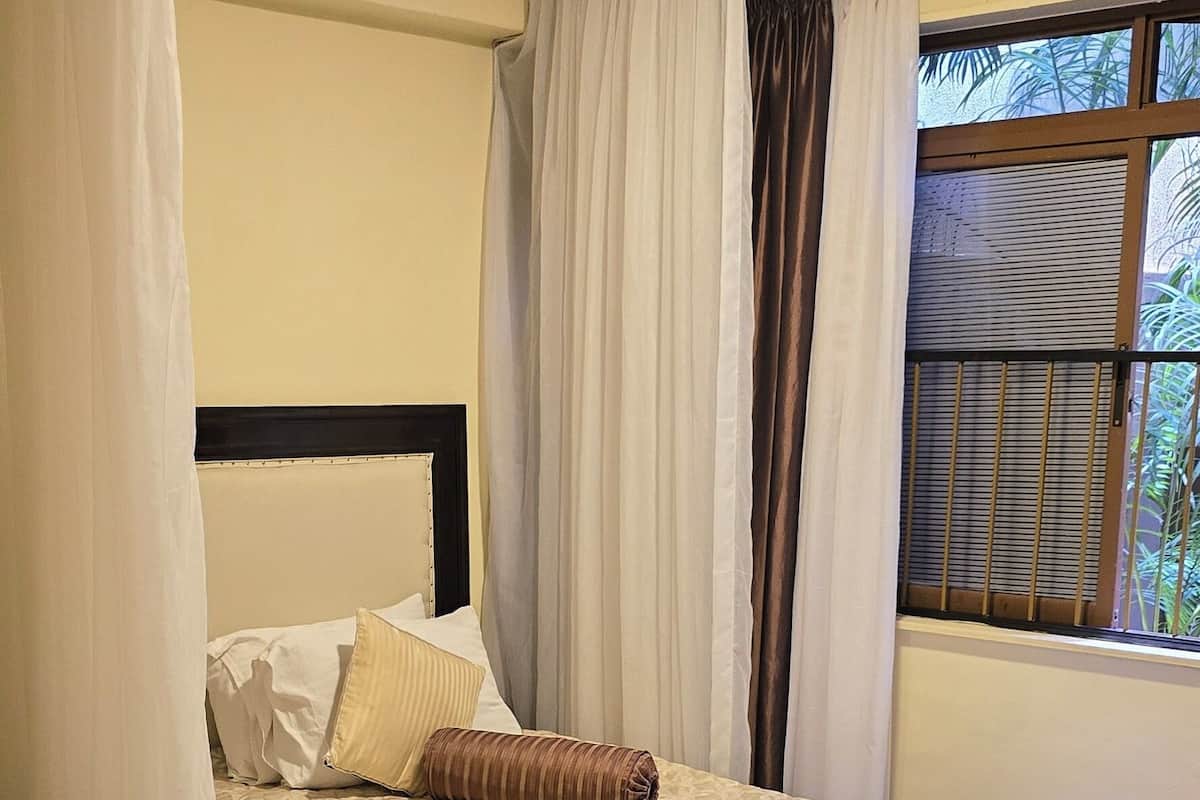 A neatly made bed with a patterned bedspread, a headboard, and a window with a view of greenery.
