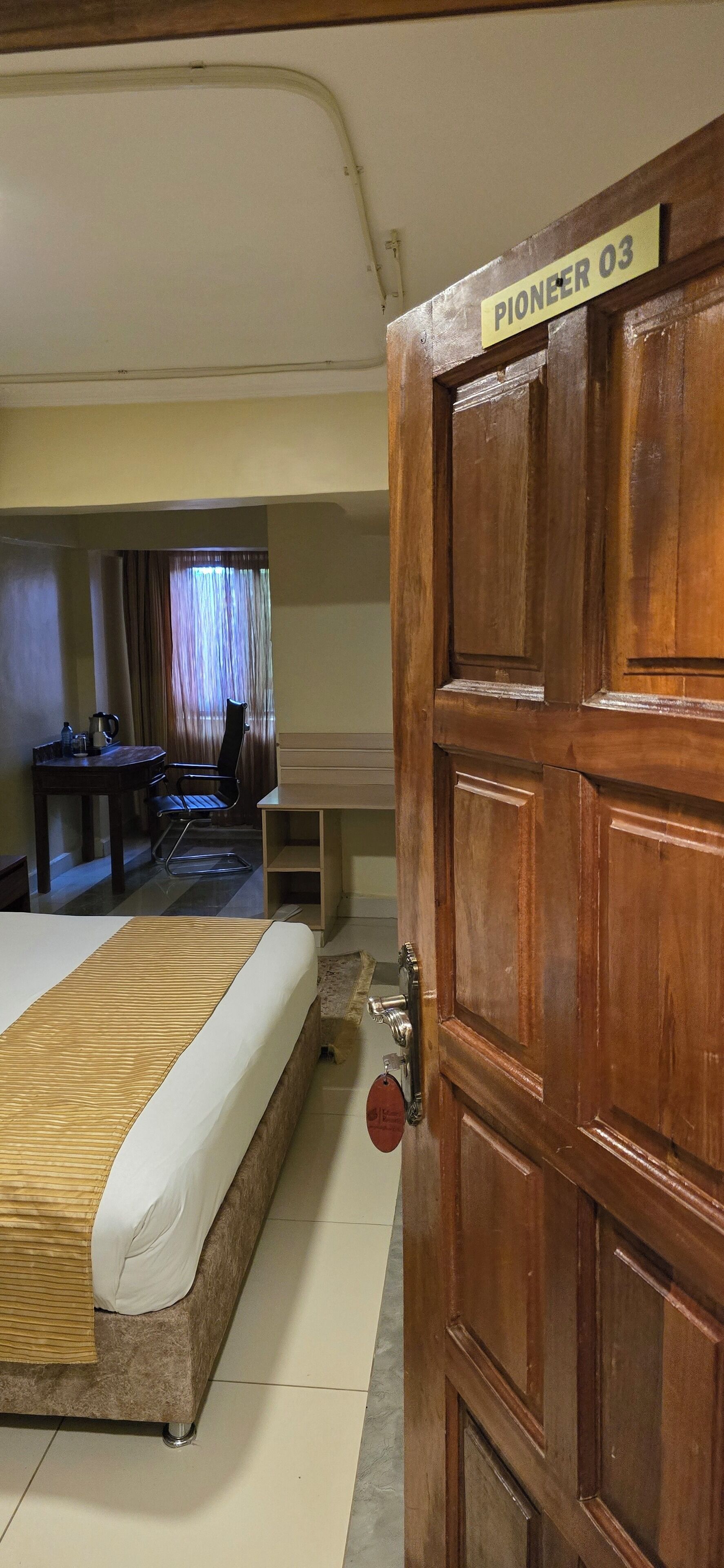 Una camera d'albergo con un letto, una porta in legno con la scritta "Pioneer 03", una scrivania con una sedia e una finestra con tende.