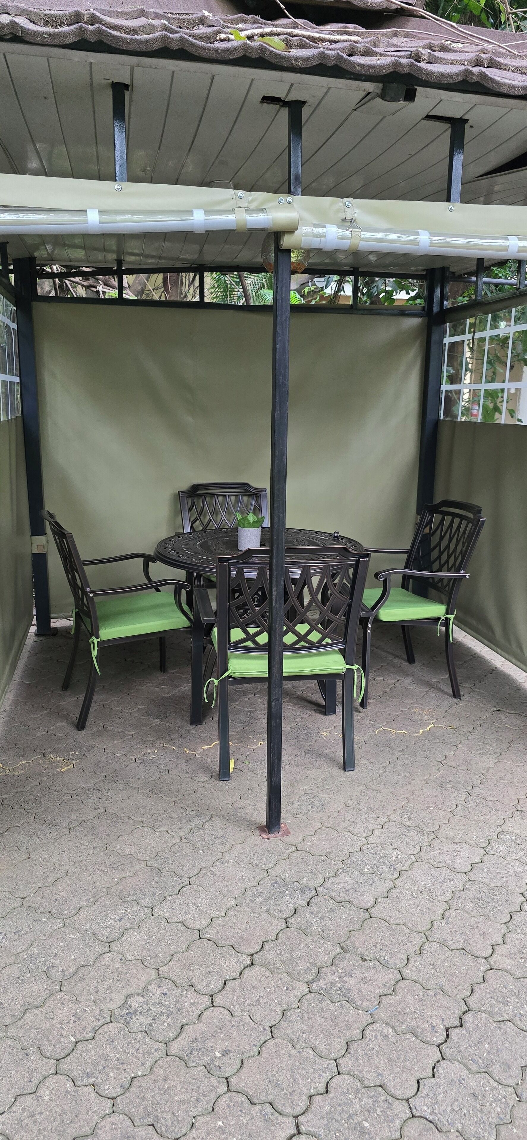 A covered patio area with a round table and four chairs, featuring a green cushioned seat and backrest.