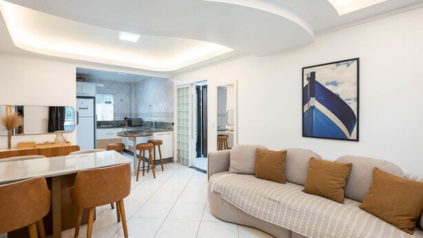 A modern living area with a sofa, dining table, and chairs. There is a kitchen in the background with a view of the outside. - Apts in Condominium 40 Meters From Bombinhas Beach - Bdss (Bombinhas)