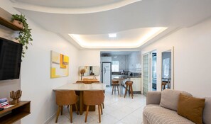 A modern living room with a sofa, a dining area with a table and chairs, a kitchen in the background, and a TV mounted on the wall. - Apts in Condominium 40 Meters From Bombinhas Beach - Bdss (Bombinhas)