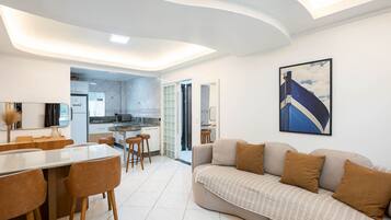 Uma sala de estar moderna com sofá, mesa de jantar e cadeiras. Cozinha visível ao fundo, com vista para o exterior.