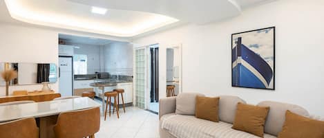 A modern living area with a sofa, dining table, and chairs. There is a kitchen in the background with a view of the outside.