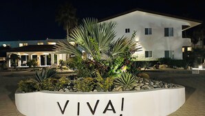 Área externa bem iluminada com uma placa que diz 'VIVA!' rodeada por plantas e um edifício ao fundo.
