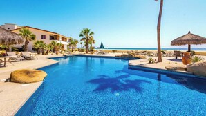 Piscina azul límpida com espreguiçadeiras e palmeiras, praia ao fundo, e um edifício com telhado de palha.