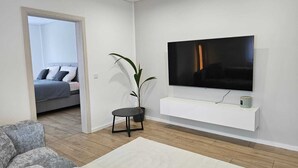 A modern living room with a flat-screen TV mounted on a white wall, a grey sofa, a small round table, and a potted plant. - Modern apartment in the heart of Ramstein (Ramstein-Miesenbach)