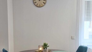 A dining area with a round table, two blue chairs, a wall clock, and a window with sheer curtains. - Modern apartment in the heart of Ramstein (Ramstein-Miesenbach)