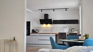 A modern kitchen with white cabinets, a black range hood, and a marble backsplash. There is a dining area with blue chairs and a small table. - Modern apartment in the heart of Ramstein (Ramstein-Miesenbach)