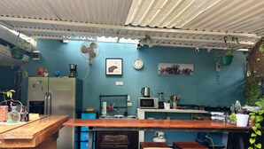 A compact kitchen with a wooden counter, a refrigerator, a microwave, and a gas stove. There are three stools and a hanging plant. - The Backside House  - Women Only - (Tamarindo)