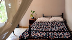 A bed with a patterned bedspread, a bedside plant, and a window with a view of greenery. - The Backside House  - Women Only - (Tamarindo)