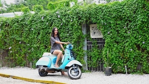 A person riding a blue scooter on a street with a green ivy-covered wall and a sign that reads "BACKSIDE" in the background. - The Backside House  - Women Only - (Tamarindo)