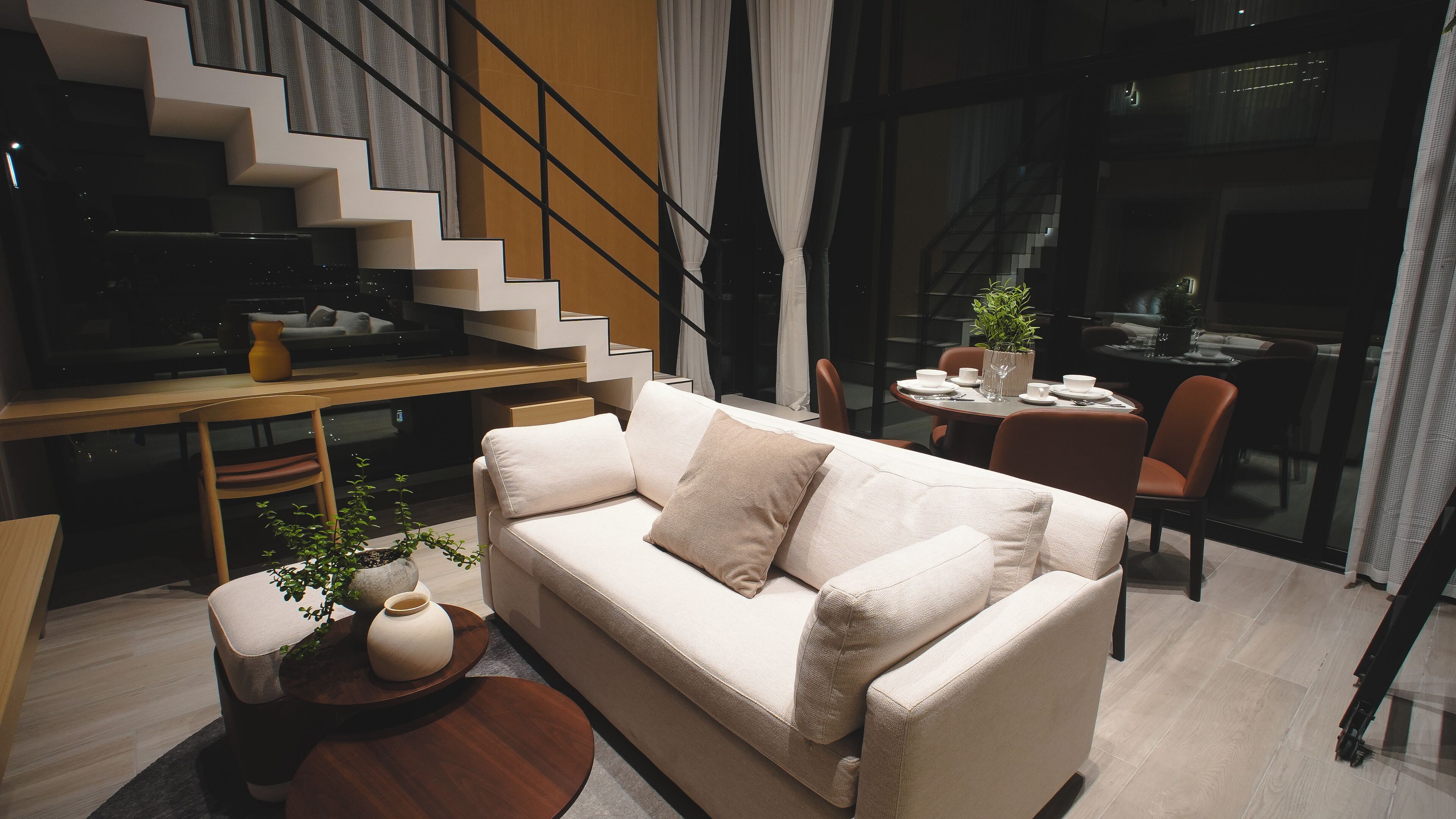 Un salon moderne avec un canapé, une table basse et un coin repas comprenant une table et des chaises. On aperçoit un escalier au fond.