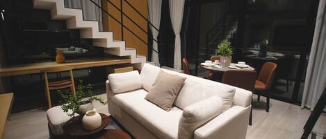 Un salon moderne avec un canapé, une table basse et un coin repas comprenant une table et des chaises. On aperçoit un escalier au fond.