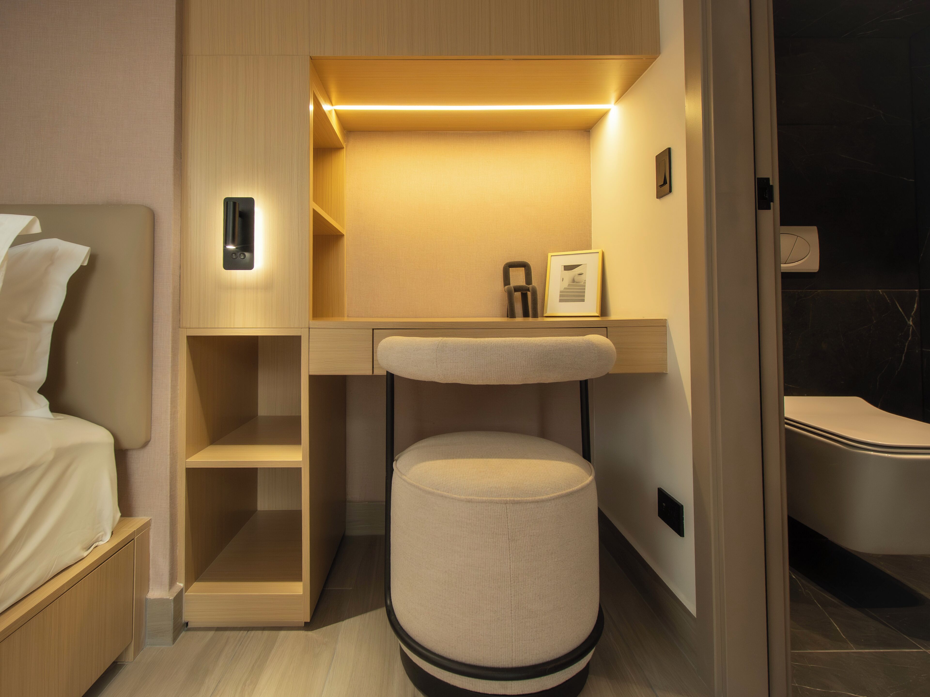 A compact hotel room with a bed, a desk with a chair, a small trash bin, and a toilet.