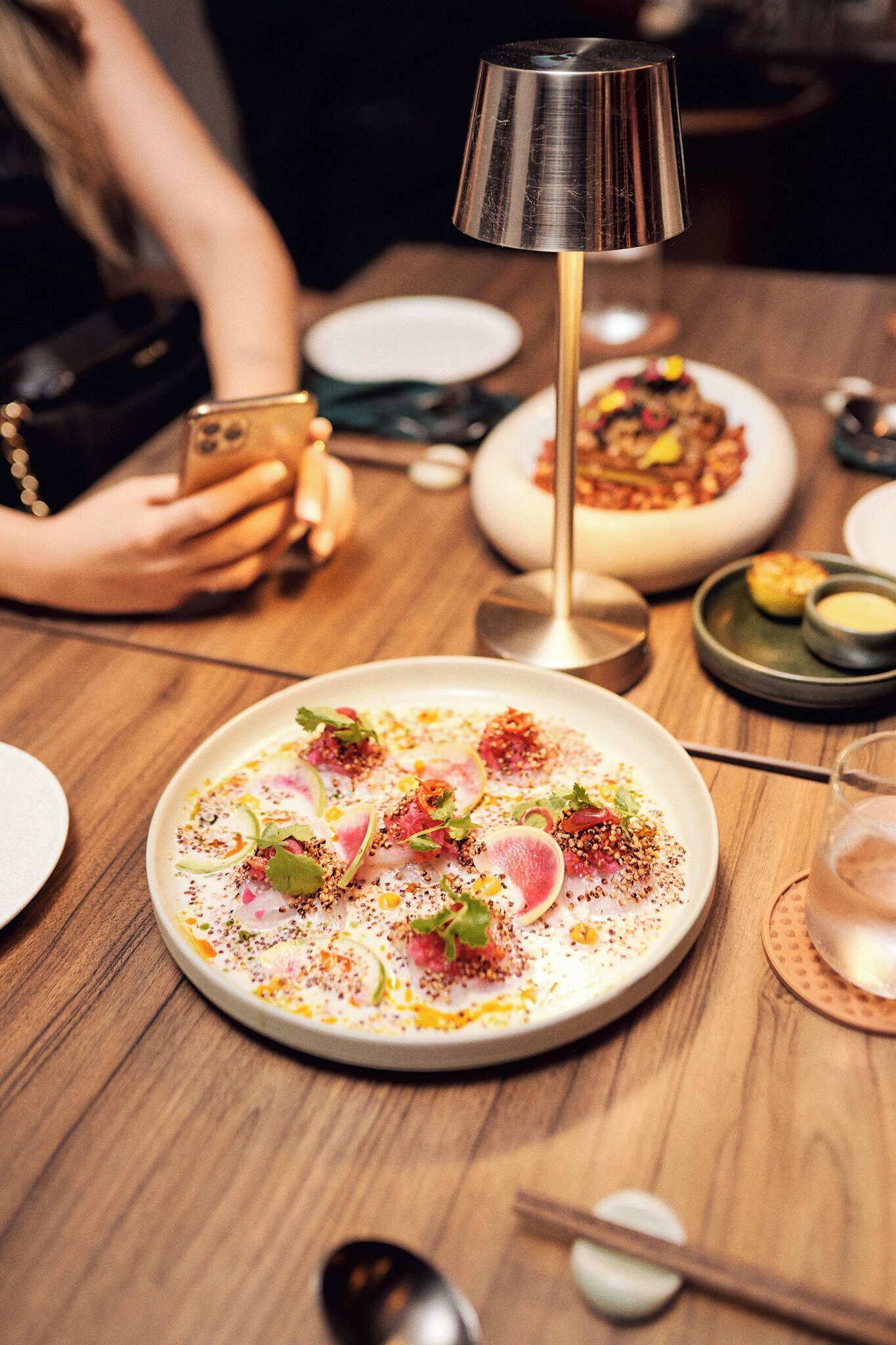 Une table à manger avec une assiette de nourriture, une lampe et une personne utilisant un téléphone intelligent.