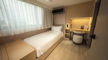 A modern hotel room with a single bed, a bedside table, a built-in bench, a small bedside table, a wall-mounted air conditioner, a bedside lamp, and a window with sheer curtains.