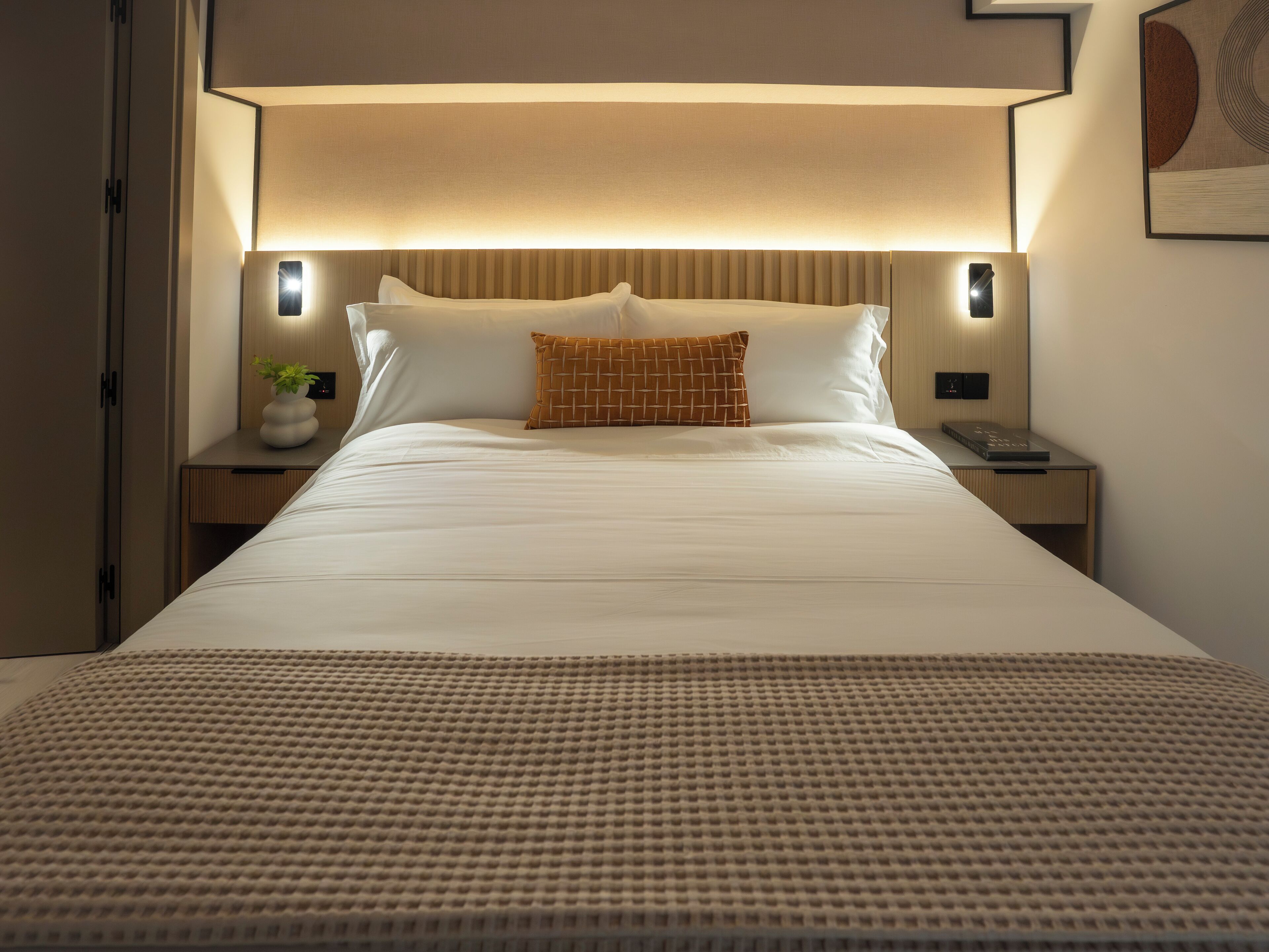 A neatly made bed with a white duvet, a brown patterned pillow, and a textured throw blanket. There are two bedside lamps and a small table with a vase of flowers.